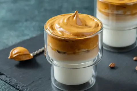 Puszyste dalgona coffee w szklance, z pianką na wierzchu i mlekiem na dole. Obok łyżeczka z pianką i ziarna kawy.