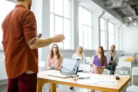 Mężczyzna z mikrofonem prezentuje swój elevator pitch grupie słuchaczy. Na stole laptop z wykresami.