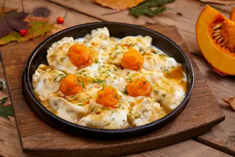 Pierożki z dynią w sosie, posypane koperkiem, podane w żeliwnej patelni na drewnianym blacie. Obok kawałek dyni.
