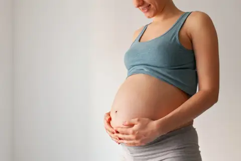 Uśmiechnięta ciężarna kobieta trzyma dłonie na brzuchu. Wkrótce może pojawić się czop śluzowy.