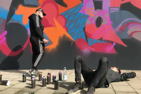 Zwei Künstler, eine Grafikdesignerin und ihr Kollege, sprühen Graffiti. Einer liegt entspannt, der andere sprüht Farbe auf die Wand.