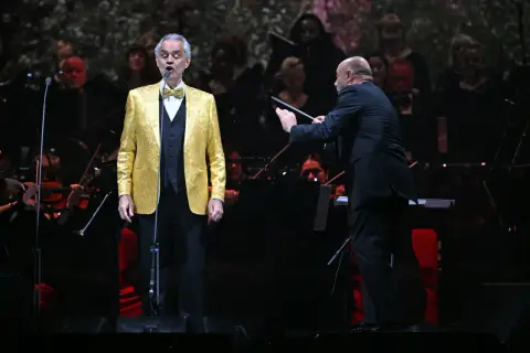 Andrea Bocelli w złotej marynarce śpiewa na scenie, dyrygent prowadzi orkiestrę. Koncert zapowiada się na niezapomniane przeżycie, jakiego można spodziewać się po Andrea Bocelli koncert 2026.