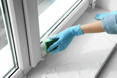 Ręka w niebieskiej rękawiczce czyści parapet gąbką. Dowiedz się, jak usunąć klej po taśmie z drzwi i innych powierzchni.