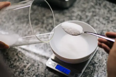 Wsypywanie cukru na łyżeczkę, która jest na wadze. Waga pokazuje 6.84 g, co pomaga ustalić, ile mg ma łyżeczka.