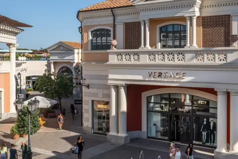 Boutiques de luxe, dont Versace, dans un centre commercial de style italien près de Rome. Des clients flânent sous le soleil.