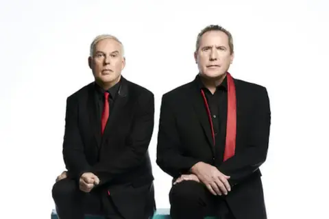 Andy McCluskey i Paul Humphreys z OMD, duet znany z niezapomnianych piosenek, w czarnych garniturach i czerwonych akcentach.