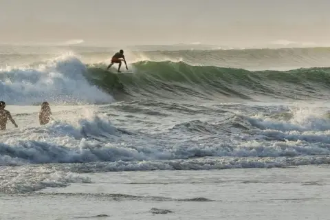 Surfer na desce ślizga się po zielonej fali, podczas gdy dwie sylwetki stoją w wodzie. To dowód, że surfing w Polsce jest możliwy!