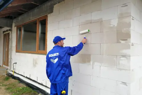 Budowlaniec w niebieskim uniformie nakłada unigrunt wałkiem na ścianę z bloczków.