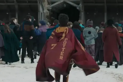 Chłopiec w pelerynie z napisem "POTTER 7" idzie przez śnieg w otoczeniu innych uczniów, jak w serialu Harry Potter.