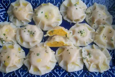 Pyszne pierogi z dynią, polane masłem i posypane szczypiorkiem, ułożone na niebieskiej paterze.