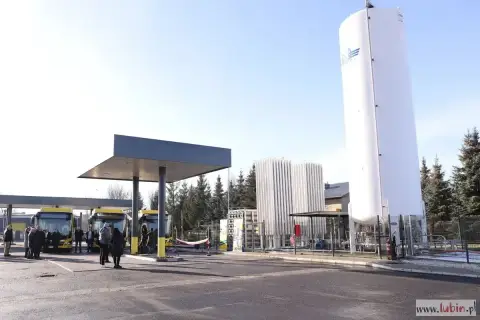 Stacja tankowania CNG. Żółte autobusy czekają pod wiatą, obok wysoki zbiornik i instalacja.