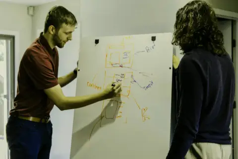 Dwóch mężczyzn planuje projekt na tablicy. Jeden pisze "content", drugi patrzy, jakby chciał uniknąć filistra.