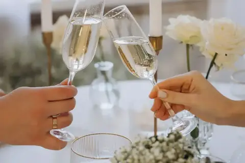 Dwie dłonie wznoszą kieliszki szampana do toastu. Elegancki stół z białymi różami i świecami. Idealny moment, by zastanowić się, ile wina na wesele potrzeba.