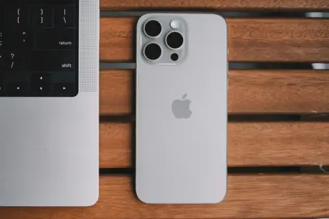 Srebrny iPhone 13 Pro z systemem iOS 26.4 obok klawiatury MacBook'a na drewnianym stole.