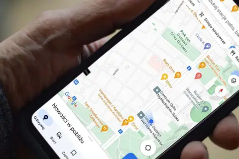 Dłoń trzyma telefon z mapą Google. Widoczna jest opcja udostępnianie lokalizacji, a także pobliskie restauracje i sklepy.