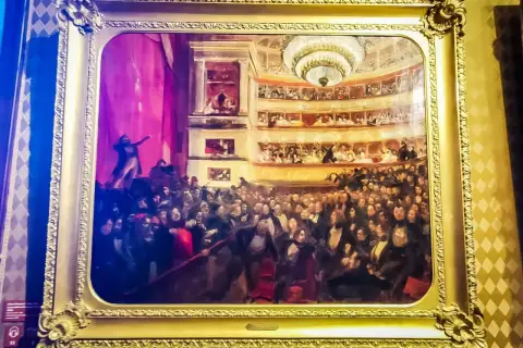 Obraz przedstawia tłum w teatrze, być może inspirowany **najważniejszymi dziełami Victora Hugo**, z widownią i lożami.