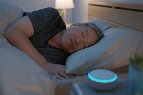 Ein Mann schläft tief und fest im Bett. Neben ihm leuchtet ein Smart Speaker, der ihm hilft, lange schlafen zu können.