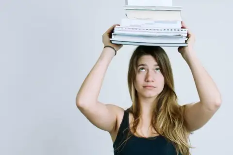 Junge Frau balanciert Bücher auf dem Kopf. Das Gewicht symbolisiert den Lernaufwand im Gymnasium und die Herausforderung des strukturierten Lernens.
