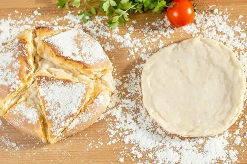 Une recette de pain maison : une miche dorée saupoudrée de farine à côté d'une pâte à pain crue, avec des tomates et du persil en arrière-plan.