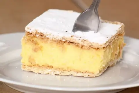 Kawałek kremowej napoleonki z cukrem pudrem, jak z przepisu babci. Widelec zagłębia się w puszysty krem.