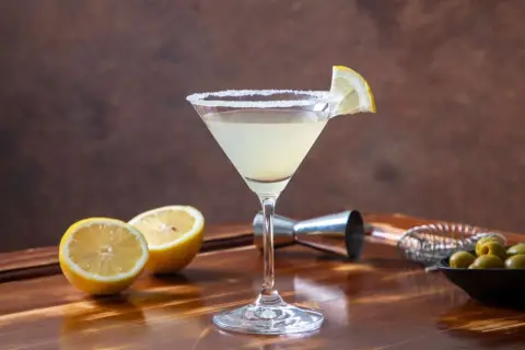 Klasyczny martini z cytryną i solą na kieliszku. W tle cytryny, oliwki i akcesoria barmańskie. Poznaj różne martini rodzaje.