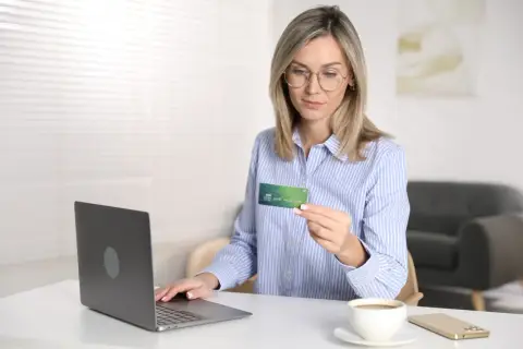 Kobieta z kartą płatniczą w ręku, gotowa do dokonania płatności online. Na biurku laptop, telefon i kawa. Reserved Paypo.