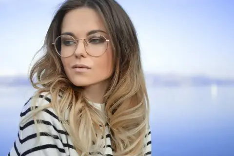 Blondynka w okularach o okrągłych oprawkach, w pasiastym swetrze, patrzy w bok na tle błękitnego nieba i wody.
