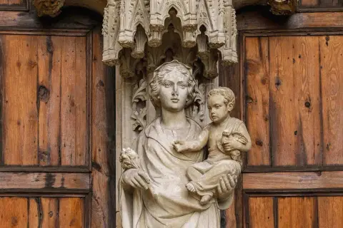 Kamienna rzeźba Matki Boskiej z Dzieciątkiem na tle drewnianych drzwi, symbolizująca teocentryzm i boską obecność w codzienności.