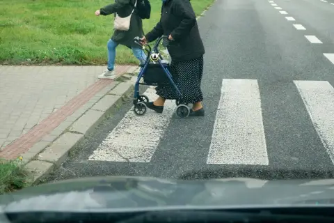 Starsza pani z chodzikiem i młodsza osoba przechodzą przez przejście dla pieszych.