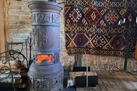 Stary, ozdobny kominek czy koza z płonącym ogniem, obok stos drewna i kolorowy kilim na ceglanej ścianie.