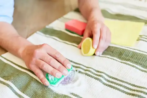 Ręce czyszczą plamę z jagód na dywanie, używając sody i cytryny.
