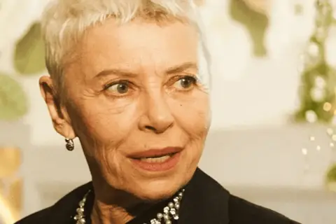 Kobieta z krótkimi siwymi włosami, w czarnej bluzce z perełkami, opowiada o tym, jak zostać aktorką.