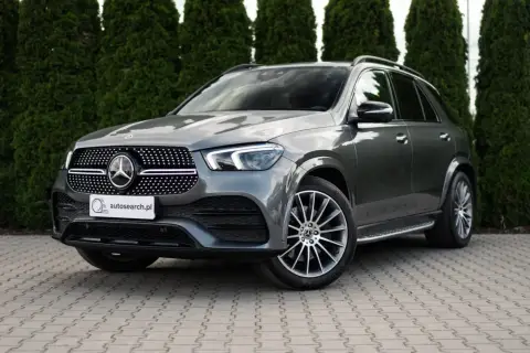 Elegancki, szary Mercedes GLE 400d SUV stoi na wybrukowanym podjeździe, otoczony gęstymi, zielonymi żywopłotami.