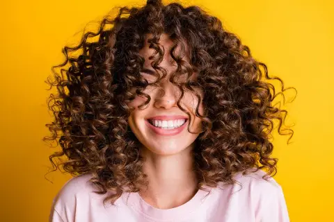Mujer sonriendo con cabello rizado castaño sobre fondo amarillo.