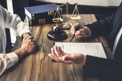 Prawnik omawia z klientem dokumenty, na stole młotek sędziowski, waga i księgi. Klient trzyma dłoń prawnika, podpisując list intencyjny.