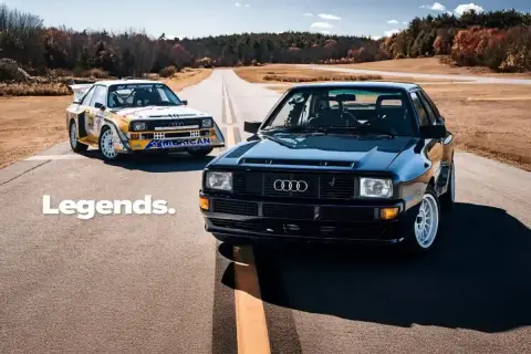 Dwa kultowe Audi S1 Quattro, jeden czarny, drugi w barwach rajdowych, stoją na pustej drodze.