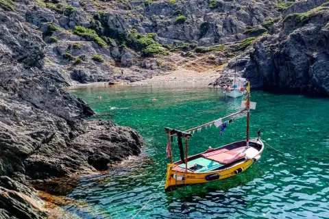 Barca amarilla en cala de aguas turquesas en el **Cabo de Creus**. Rocas escarpadas y gente en la playa al fondo.