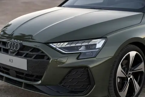 Nowe Audi A3 w zielonym kolorze prezentuje się elegancko. Widoczny fragment maski, grill z logo i reflektor podkreślają jego nowoczesny design.