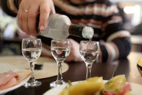 Ręka nalewa wódkę do kieliszków, tworząc potencjalny rekord promili. Na stole wędlina i przekąski.