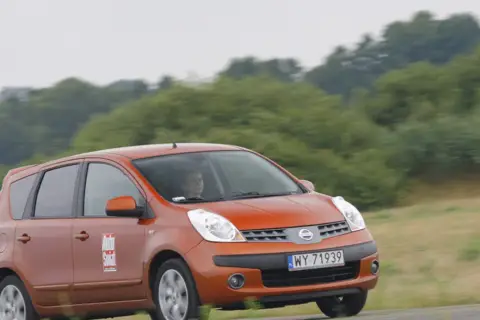 Pomarańczowy Nissan Note E12 z kobietą za kierownicą jedzie po drodze wśród zieleni.