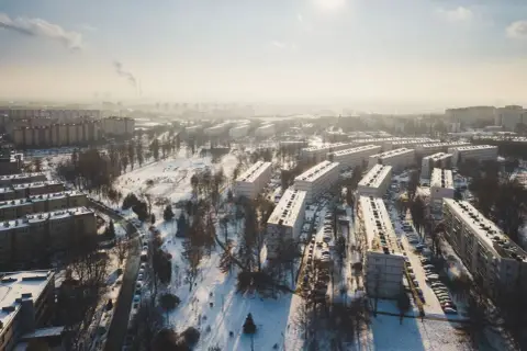 Zimowy krajobraz Krakowa z blokami mieszkalnymi i parkiem. Widać dymiące kominy w oddali, co może wpływać na ceny mieszkań w Krakowie.