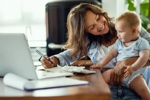 Lächelnde Mutter mit Baby auf dem Arm schreibt am Laptop. Sie plant die Zukunft und legt für das kinder sparbuch.