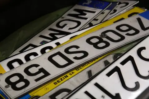 Stos **tablic rejestracyjnych państw** w różnych kolorach i z numerami. Widać fragmenty z PL, F, a także napisy "BEZIN SA MERCEDES".