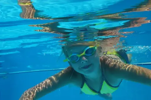 Enfant souriant sous l'eau, lunettes de piscine, maillot jaune. Attention à l'otite de piscine après ces moments joyeux.