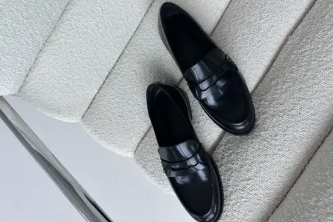 Eleganckie, czarne, lakierowane mokasyny leżą na białym, teksturowanym fotelu. Idealne, by dowiedzieć się, jak zmiękczyć skórzane buty.