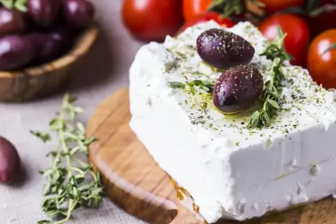 Kostka sera feta, polana oliwą i posypana ziołami, z oliwkami i pomidorkami w tle. Właściwości sera feta doceniane są w kuchni śródziemnomorskiej.