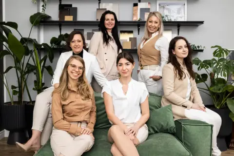 Siedem kobiet w eleganckich strojach siedzi i stoi w nowoczesnym wnętrzu, otoczone roślinami i próbkami materiałów.
