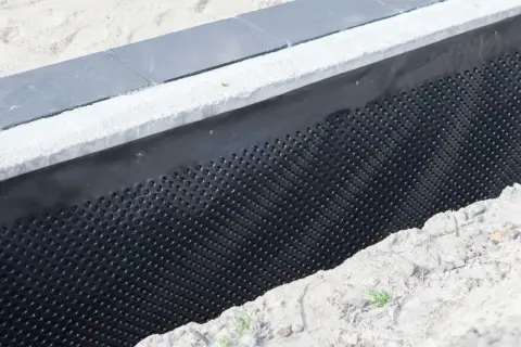 Czarna folia kubełkowa montaż zabezpiecza fundament przed wilgocią. Widać ją przy betonowym murku i piasku.