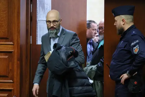 Mężczyzna w garniturze z książką i kurtką, obok policjant i inni mężczyźni w pomieszczeniu.