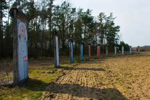 Fragment muru berlińskiego z kolorowymi malunkami, stojący na tle lasu i zaoranego pola.
